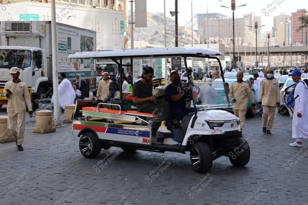 صورة تظهر مشهدًا لشارع مزدحم في مدينة سعودية. يظهر في الصورة سيارة جولف بيضاء تستخدم كإسعاف صغير, تتوسط الشارع, عليها مرتبة برتقالية ومظلة. يجلس عليها شخصان يرتديان ملابس غير رسمية ويبدو أنهما من الطاقم الطبي. من خلفهم, تسير مجموعة من الأشخاص يرتدون زي الإحرام الأبيض والزي الرسمي. تظهر أيضًا شاحنة بيضاء في الخلفية مع كتابات بالعربية. توجد مبانٍ متعددة الطوابق ومدن حديثة خلفهم, مما يشير إلى مدينة حضرية مزدحمة.