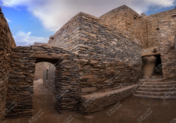 An image of a narrow street between old stone buildings that appear to be historical ruins. The walls are made of beige and brown stones, and in the middle of the image, there is an arched entrance leading to a narrow path. On the right side, there is a stone staircase leading to a higher level. The sky is blue with light clouds in the background.