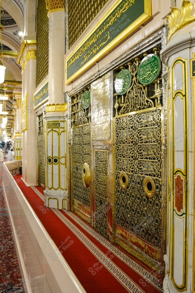 Image of an interior scene in the Prophet\'s Mosque in Medina, Saudi Arabia. The photo shows the arcade facing the Prophet\'s Chamber, which is adorned with Islamic calligraphy and Quranic verses on the ornate gate. The floor is covered with a red patterned carpet. Lighting is provided by lamps hanging from the ceiling.