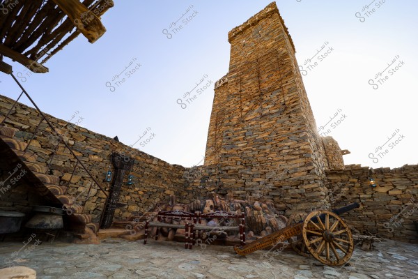 The image shows a traditional old stone building with a tall, rectangular shape resembling a tower, surrounded by thick stone walls. Stone stairs on the left lead to the roof. Along the walls, traditional blue lanterns embellish the scene. In the foreground, there is an old wooden cannon, and the setting appears to be a historic courtyard of an ancient place.