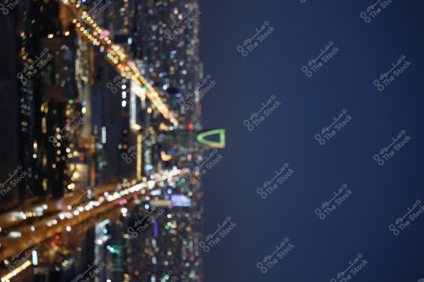 A nighttime image of Riyadh in Saudi Arabia showing an illuminated cityscape. The Kingdom Tower, with its iconic architectural shape, is centrally featured, surrounded by other brightly lit buildings and busy roads filled with cars. The sky is clear and dark.