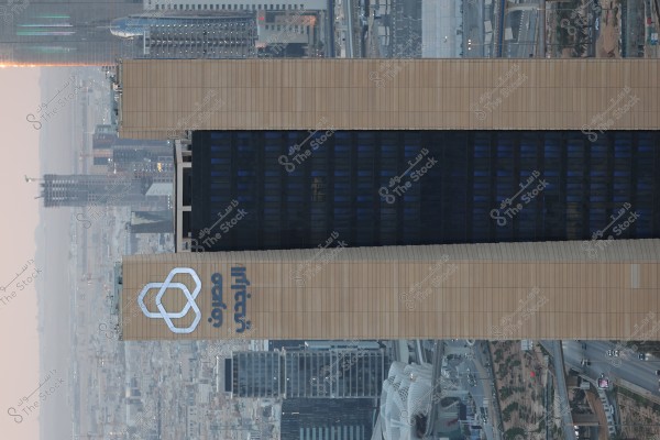 صورة لبرج طويل يظهر بشكل بارز في مشهد المدينة. البرج مغطى بواجهة زجاجية سوداء ولون بني داكن مع شعار وكتابة بارزة عليه. يحيط بالبرج مباني شاهقة أخرى وجسور وطرق, مما يشير إلى منظر حضري متطور. الصورة تبدو وكأنها مأخوذة من ارتفاع يسلط الضوء على البنية المعمارية الحديثة للمدينة.