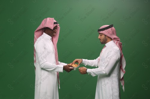 لقطة مقربة لرجل سعودي يرتدي الزي السعودي التقليدي يحمل طبق من المعمول, إفطار صائم, فطور صحي, ضيافه , معمول من التمر, خلفيه خضراء.