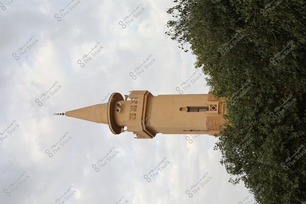 An image of a mosque\'s minaret standing tall against a cloudy sky, with dense green trees beside it. The overcast sky adds a serene atmosphere to the scene. The design of the minaret showcases traditional Islamic architectural style.