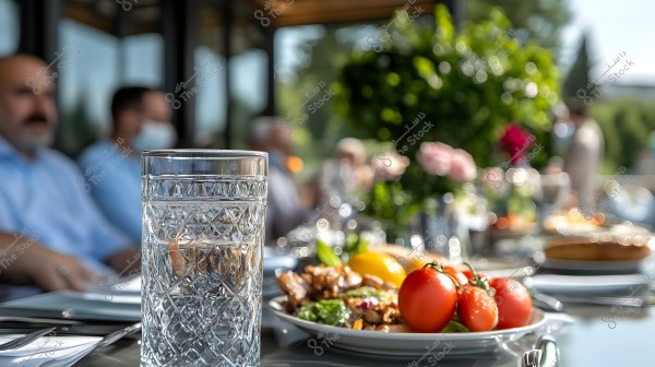 The image shows a crystal glass of water placed on a table set with dishes containing vegetables, including tomatoes and lemon. In the background, people are sitting around the table, with an outdoor setting filled with greenery and flowers.