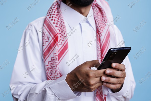 An image of a man wearing a white thobe and a red and white keffiyeh draped over his shoulders, using a black smartphone. The attire is traditional Gulf style. The background is light blue.