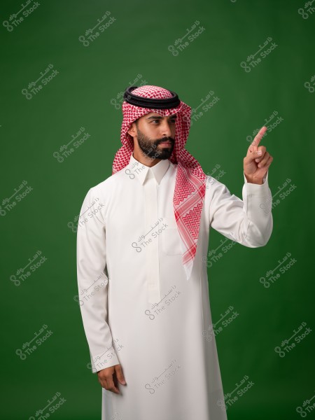 A man wearing traditional Saudi attire, featuring a white thobe with a red and white checkered shemagh and an agal. He stands against a green background, pointing upwards with his finger.