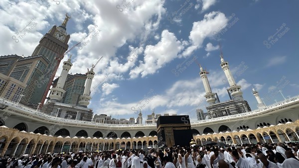 Image of the Kaaba and Al-Masjid Al-Haram in Mecca, with crowds of people performing Tawaf around the Kaaba. The Clock Tower and surrounding architecture are visible under a cloudy sky.