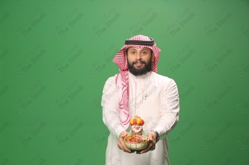 شاب سعودي على خلفية خضراء يرتدي الثوب و الشماغ، يقوم بعمل حركات بيده،يمسك بسلة الحلوى و يقدمها، احتفالات العيد