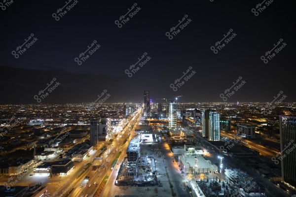A night view of a large city featuring modern buildings and illuminated skyscrapers. Wide roads stretch across the city with visible traffic. The dark sky adds to the beauty of the nighttime scene.