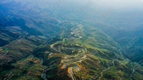 An aerial view of a winding mountain road stretching through a green mountain range. The road twists dynamically between the hills, surrounded by scattered green vegetation. The horizon appears misty, adding a serene and impressive dimension to the natural landscape.