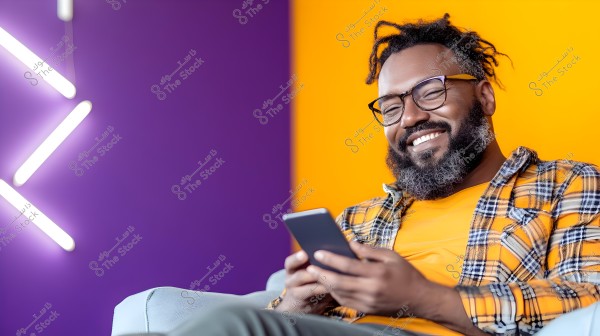 A portrait of a smiling man with a beard and glasses, sitting on a sofa holding a smartphone. He is wearing an orange shirt under a plaid overshirt. The background is brightly colored with purple and orange, featuring white neon lights on the left side.