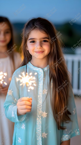 A portrait of a young girl wearing a blue dress adorned with white stars, holding a lit sparkler. In the background, another girl is out of focus, wearing a light-colored dress and holding a lit sparkler as well. The scene appears to be a celebration outdoors at dusk, with a natural backdrop.