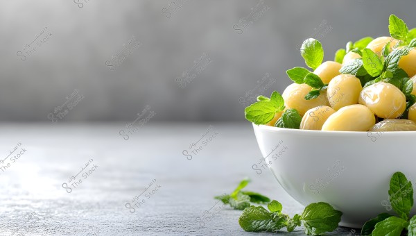 The image shows a white bowl containing large yellow olives or lupini beans with green mint leaves. The bowl is placed on a gray surface with mint leaves scattered around it. The background is smooth and blurred with gray tones.
