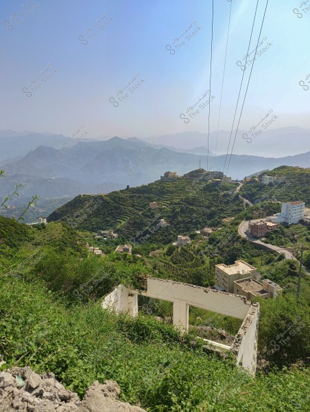A scenic view of green mountains interspersed with roads and power lines, with several buildings scattered among the mountainous terrain. The horizon features a range of mountains under a clear blue sky with light mist.