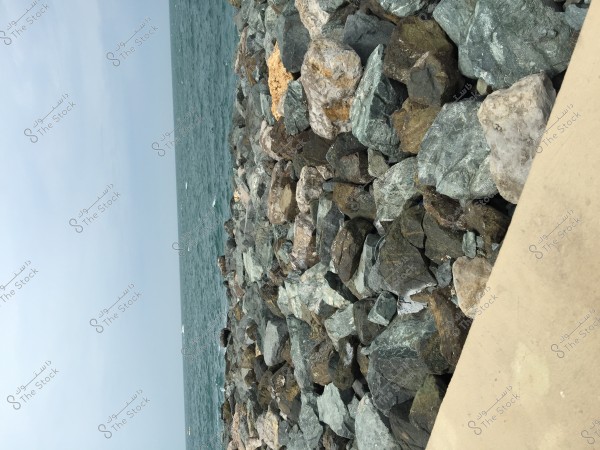 An image of a seaside area with a collection of large rocks in various colors and textures, stacked next to each other. The blue water is visible in the background under a cloudy sky.