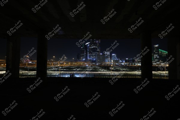 A nighttime cityscape illuminated with neon lights, featuring tall skyscrapers in the background. The city is interwoven with illuminated bridges, and a partially lit ceiling is visible at the top, adding a dramatic tone to the scene.