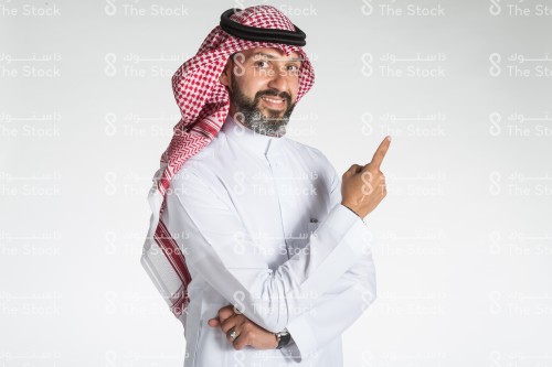 بورتريه رجل سعودي يقف على خلفية بيضاء، يرتدي الزي السعودي الرسمي،ينظر الى الكاميرا و يشير باصبعه السبابه نحو شيء معين