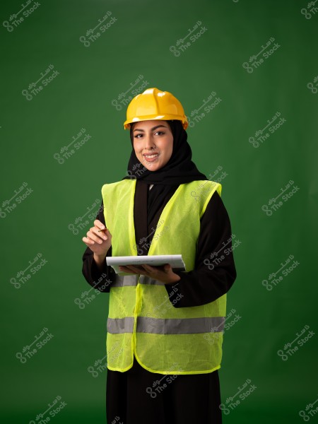 Image of a woman wearing a black hijab, a yellow safety helmet, and a bright yellow safety vest. She stands against a green background, holding a notebook, smiling, and appears to be taking notes. The portrait might be from the Gulf region based on the clothing style.