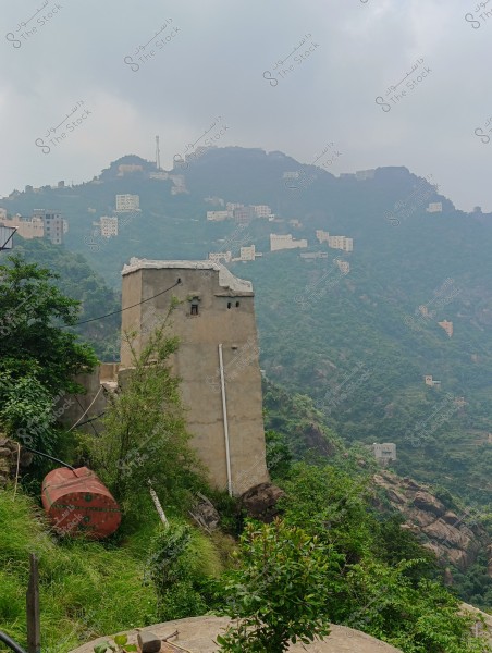 An image of a scenic view in a mountainous green area. In the foreground, there is a small water tower next to a red metal tank. The background consists of mountains covered in greenery with scattered white houses on the slopes. Clouds partially cover the sky.
