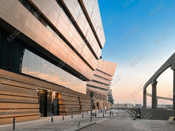 The image shows a modern-designed building with a glass facade reflecting the sky at sunset. The building features horizontal lines and a distinctive geometric shape. To the right, there is a straight concrete bridge extending into the distance. The sky is clear, with a blue color transitioning to orange at the horizon, creating a calm and beautiful atmosphere.