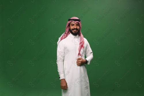 رجل سعودي يقف على خلفية خضراء يرتدي الثوب و الشماغ، يقوم بمسك الشماغ وهو مبتسم ، احتفالات العيد