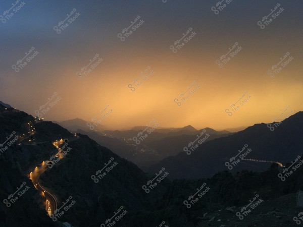 A natural landscape of a mountain under an orange evening sky with warm lighting. A winding road lit with orange lights extends through the mountains in the foreground, creating an engaging view. The horizon features hills and mountains that enhance the natural beauty of the scene.