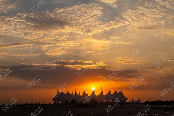 An image of a sunset behind large, polygonal tent-like structures with a unique artistic design. The sky is scattered with clouds, and the orange and yellow hues fill the horizon, adding beauty and tranquility to the scene.