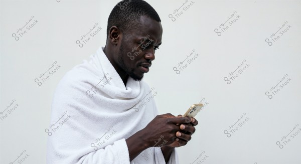 An image of a man wearing a white garment using a smartphone. The man is pictured from the side, looking at the phone with focus and attention. The background is white and plain.