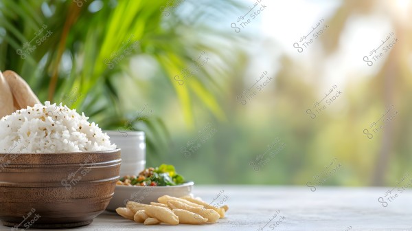 An image showing a wooden bowl filled with cooked white rice, accompanied by a small dish of chopped vegetables. There are also crispy snack pieces on the table. The background features green plants, adding a sense of calm and natural beauty.