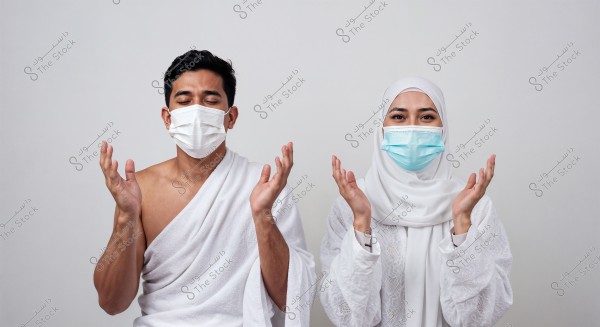 An image of a woman and a man wearing Hajj attire with medical face masks. The woman is wearing a white hijab and an embroidered caftan, while the man is dressed in a traditional white ihram. Both are raising their hands in a prayer position.