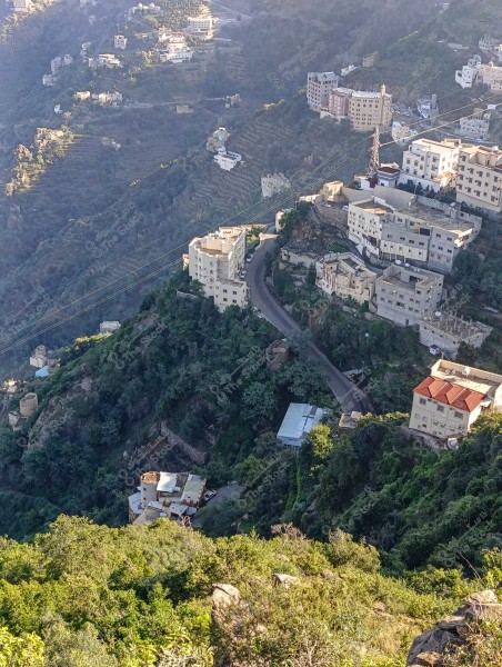 An image of a mountainous landscape featuring modern buildings scattered along terraced hillsides. Winding roads intersect the mountains, with dense greenery below. Natural lighting highlights the details of the buildings and the lush environment.