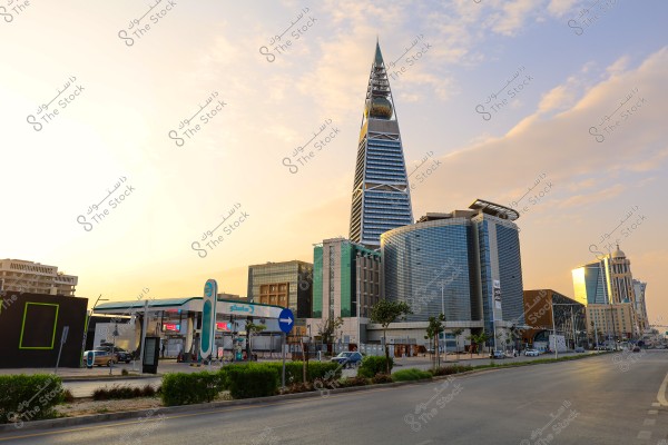 The image shows an urban scene in Riyadh, Saudi Arabia, featuring the distinctive Al Faisaliyah Tower. The tower is tall with a pyramid shape and topped with a glass orb. Surrounding the tower are modern buildings, including a round glass office building and a gas station. The sky is bright with sunlight in the background.