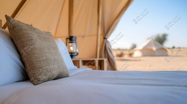 Image of the interior of a luxury tent in the desert showing a bed with a brown woven pillow and a bedside lantern. In the background, another tent is visible under a clear blue sky with sparse trees on the horizon.