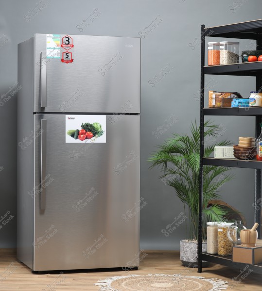The image shows a silver refrigerator with stickers detailing warranty information. On the fridge door, there\'s a sticker featuring a variety of vegetables, including cucumber, tomatoes, lettuce, broccoli, and onion. Next to the refrigerator is a metal shelf holding a variety of packaged and canned foods such as rice, pasta, and tomatoes. A small circular rug is on the floor, and there’s a green plant in the corner of the room.