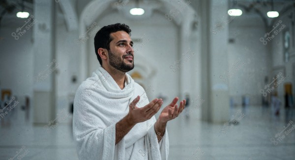Image of a man wearing white ihram clothing inside a mosque. He appears focused and contemplative as he raises his hands in prayer. The background shows architectural details of the mosque with arched decorations and hanging lights.