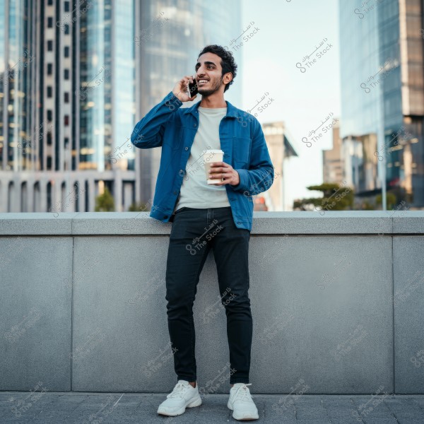 A photo of a man standing in an urban area in front of tall buildings, talking on the phone with a smile and holding a coffee cup. He is wearing a blue jacket, a white shirt, dark pants, and white sneakers.