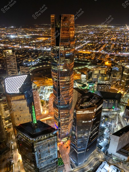 Aerial night view of a city with towering illuminated buildings, featuring a tall tower lit with orange lights in the center. The city appears expansive with city lights stretching to the horizon in the background.