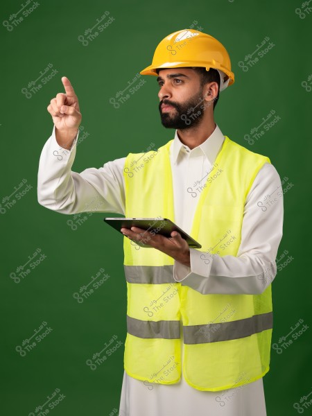 Image of a man wearing a yellow safety helmet and a high-visibility vest over a white robe, holding a tablet in one hand and pointing with the other hand, set against a green background.