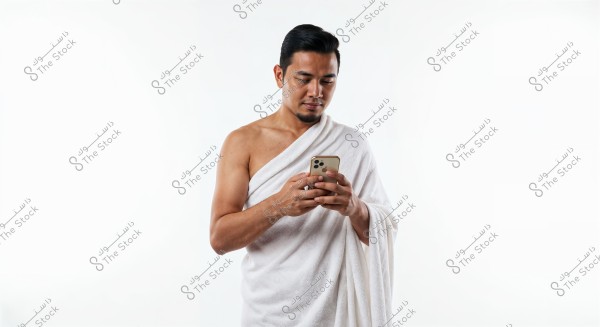 A portrait of a man wearing a white wrap, looking at a mobile phone in his hands. He appears to be wearing traditional Ihram clothing used in Hajj or Umrah in Islam. The background is completely white.