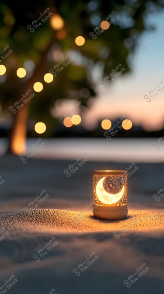 A lit lantern with a crescent design on the sand, with a background of twinkling lights in a peaceful night setting, under a sunset sky.