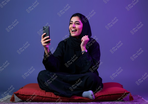 An image of a woman sitting on a large red cushion, wearing a black abaya and a black hijab, holding a smartphone in her right hand and smiling broadly. The lighting is dim with a purple hue and she is seated on a carpet. The background is a solid purple color.