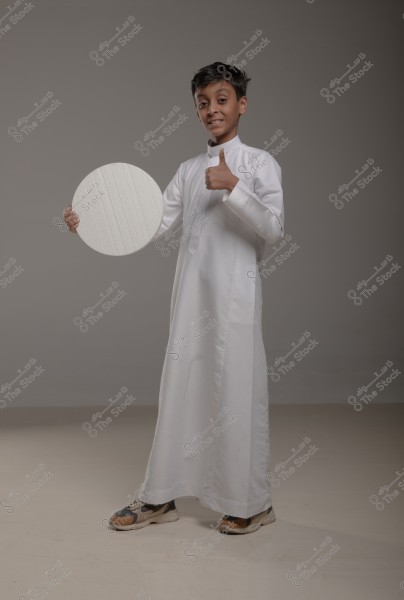 The image shows a boy wearing a traditional white thobe, holding a white circular board in his right hand. The boy gives a thumbs-up gesture, standing on a gray floor in a photography studio.