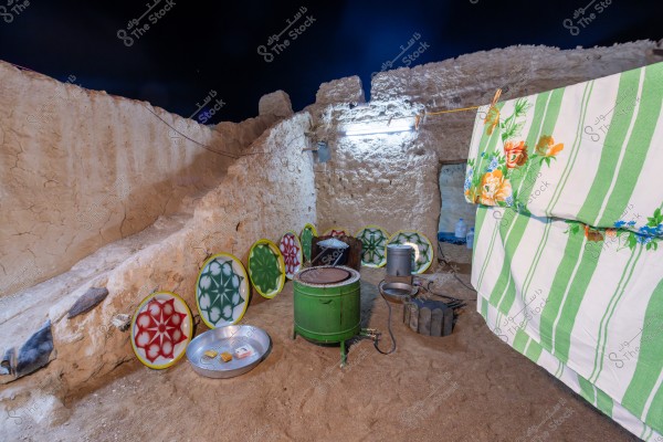An image showing a traditional outdoor courtyard with mud walls under a night sky. There are old metal cooking pots, a stove, and a sandy ground. A green cover with orange flowers and green and white stripes is hung up. Metal trays are leaning against the wall.
