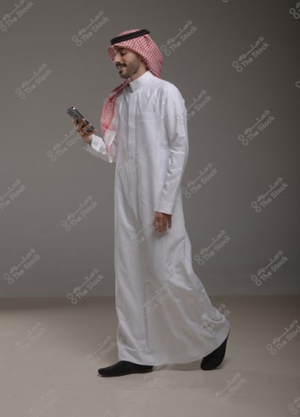 Image of a man wearing traditional Arabic attire, a thobe with a shemagh and agal, holding a smartphone and smiling while walking. The background is gray, and the setting appears professional and simple.
