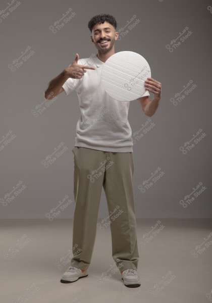 A portrait of a man standing in a studio. He is wearing a white shirt and beige pants, smiling and holding a white circle. His features suggest he might be of Middle Eastern descent.