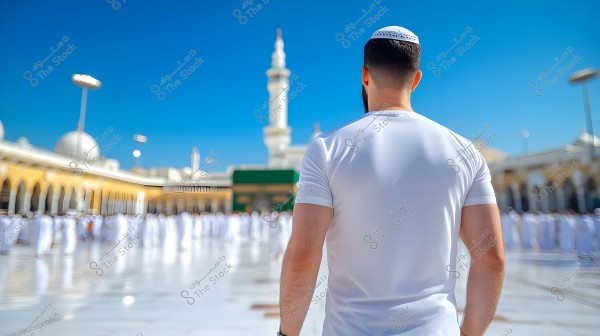 Rear view of a man wearing a white shirt and a small head covering, standing in the courtyard of the Prophet’s Mosque in Medina, Saudi Arabia. In the background, there is a white minaret and a building with white domes, with a large gathering of people dressed in traditional white clothing.