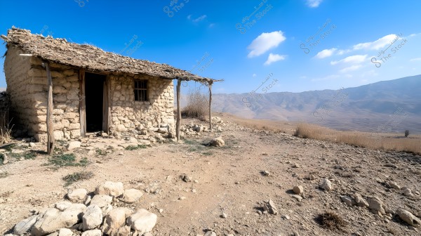 A simple stone hut situated in a dry area, featuring a thatched roof and scattered vegetation around it. The hut overlooks a mountainous landscape with a clear blue sky and a few white clouds.