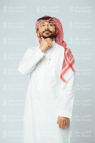 بورتريه رجل سعودي مبتسم يرتدي الزي السعودي التقليدي ويضع يده تحت دقنه بينما ينظر للأعلى وهو يفكر، شاب يتأمل