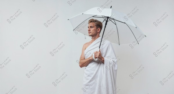 The image shows a man standing against a white background holding a white umbrella. The man is draped in a white cloth covering his left shoulder and parts of his body, creating an appearance similar to traditional attire or a towel. The pose and composition give the image a contemplative and balanced feel.
